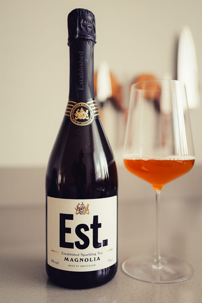 Fles Est. Sparkling Tea Magnolia met zwarte folie op de hals naast een glas amberkleurige bruisende thee