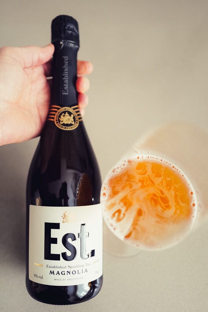 Een hand houdt een fles Est. Sparkling Tea Magnolia vast naast een gekanteld glas oranje bruisende thee