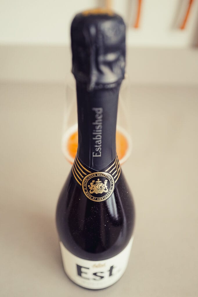 Flessenhals van Est. Sparkling Tea met zwarte folie en gouden zegel, met een glas bruisende thee onscherp erachter