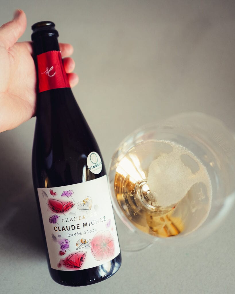 Hand houdt fles Claude Michez Cuvée Flore champagne vast naast een glas met goudgele champagne