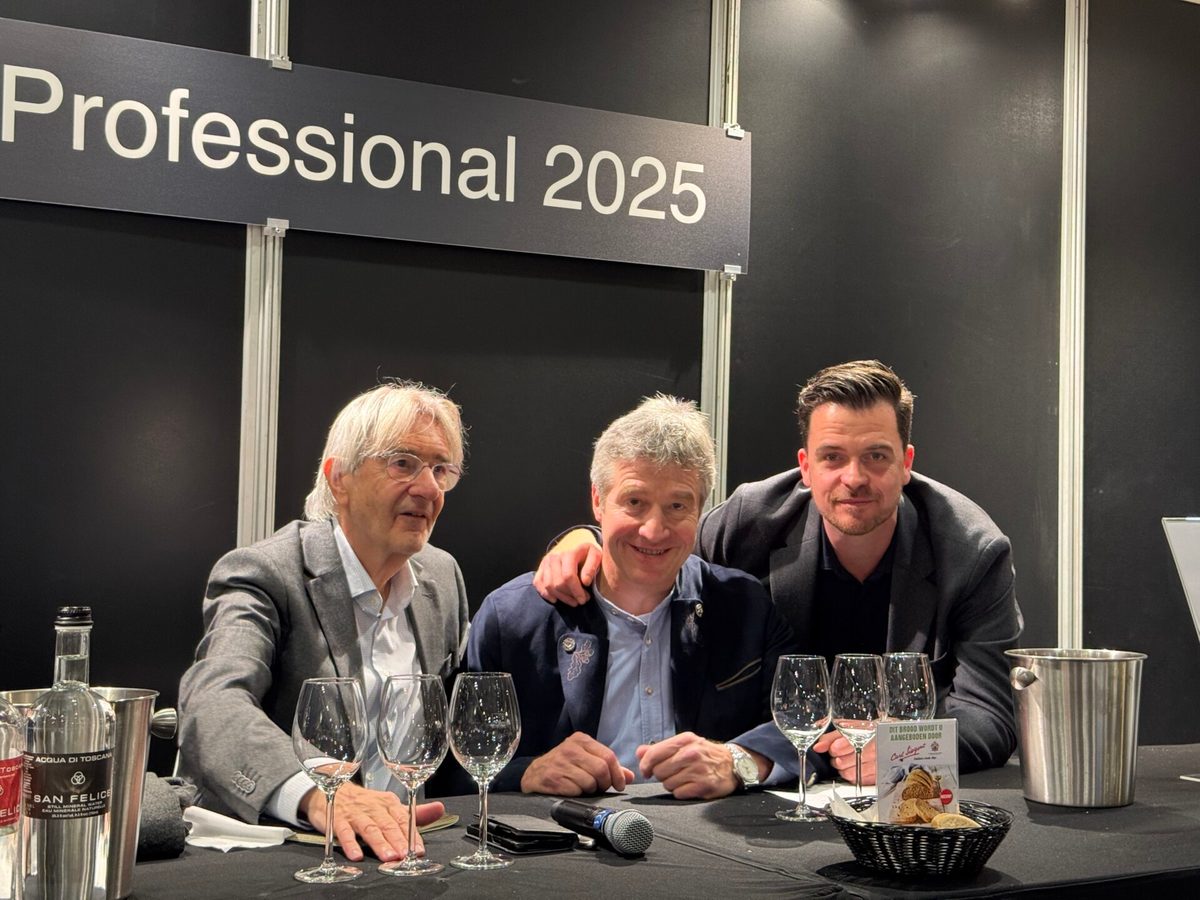 Drie mannen poseren samen aan een proeftafel met wijnglazen onder een bord Professional 2025