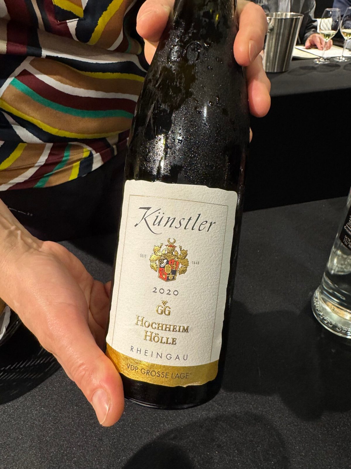 Fles Künstler 2020 GG Hochheim Hölle Riesling met wapen-etiket, vastgehouden boven een donkere tafel