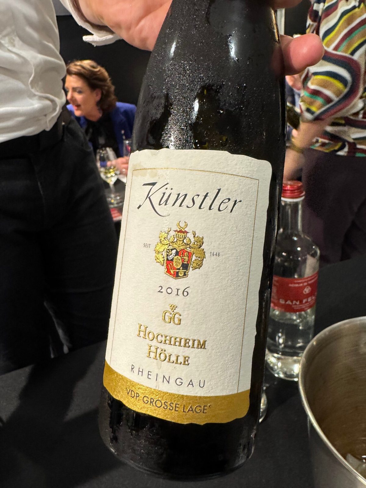 Fles Künstler 2016 GG Hochheim Hölle Riesling met condens, vastgehouden naast een gestreepte mouw