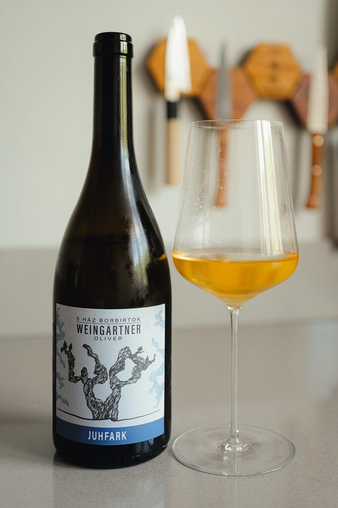 Fles Weingartner Oliver Juhfark naast glas amberkleurige oranjewijn op aanrecht
