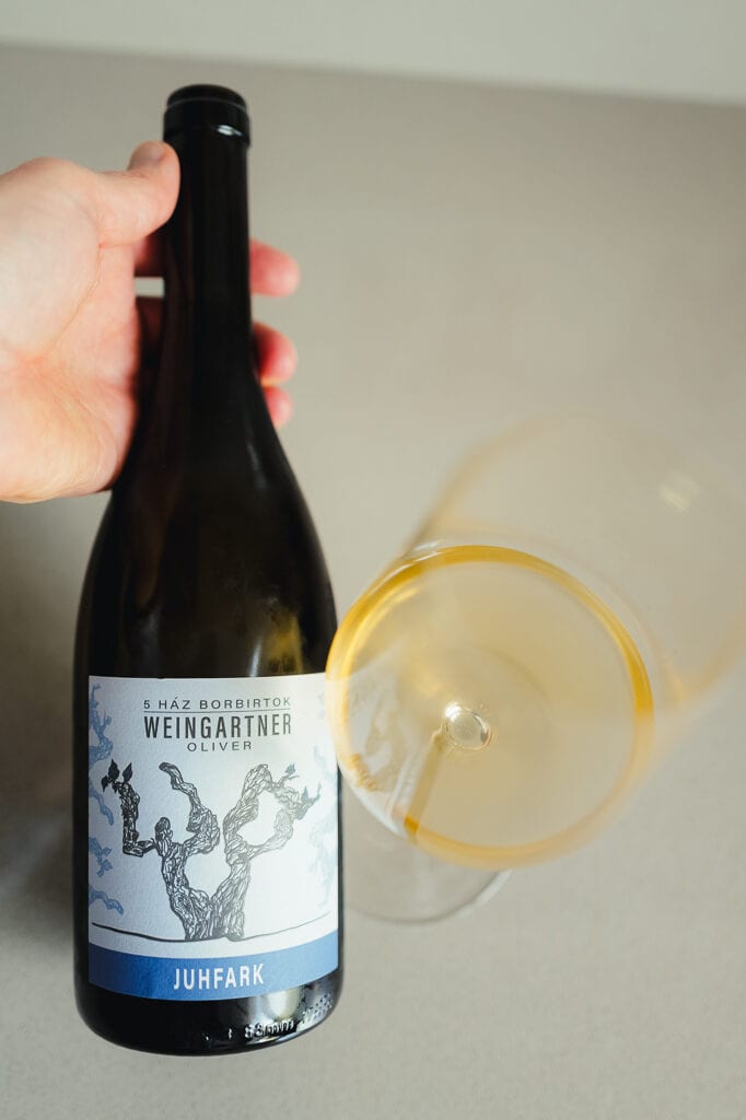 Hand houdt fles Weingartner Oliver Juhfark schuin naast liggend glas oranjewijn