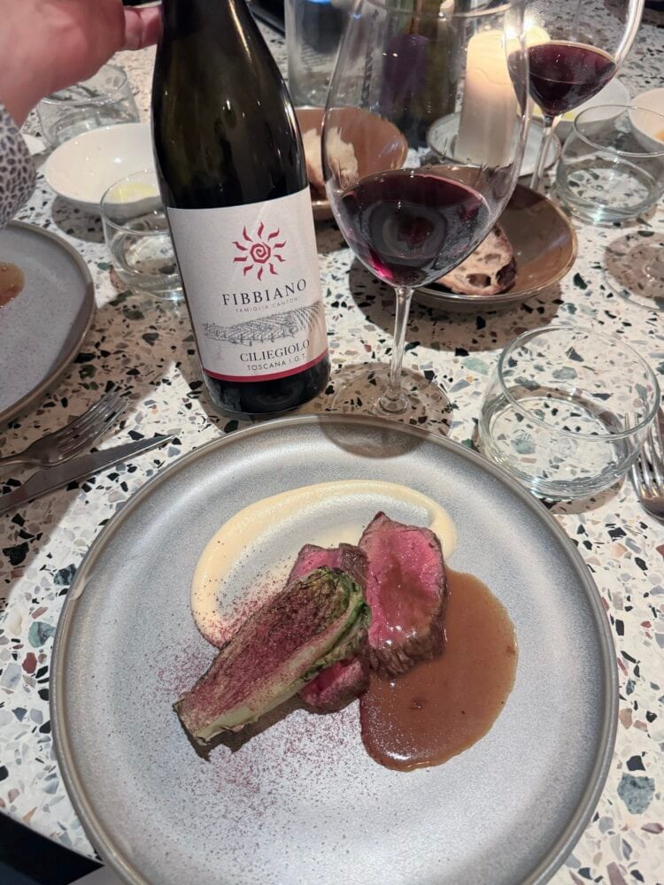 Fibbiano Ciliegiolo Toscana red wine bottle next to a plate of pink seared meat in sauce and a glass of red