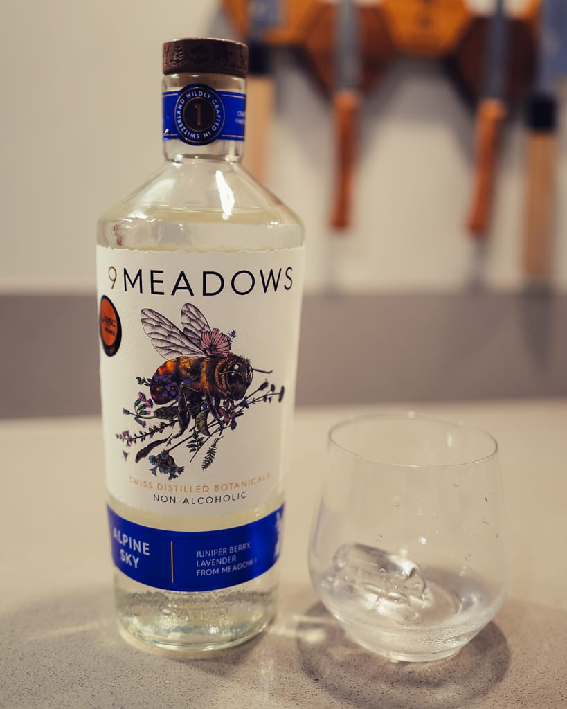 Fles 9 Meadows Alpine Sky naast een leeg glas met ijs op een lichte tafel