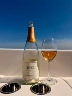 Fles Wijndomein De Boe Rosé Brut 2023 naast een glas koperkleurige mousserende wijn op een boot, met het water van de Oosterschelde erachter
