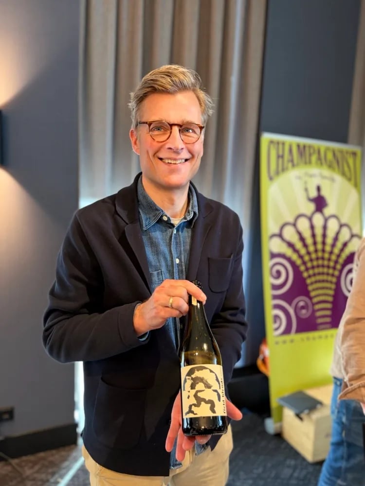 Mark Haasdijk of Champagnist holding a Field Blends bottle in front of the yellow-purple Champagnist banner