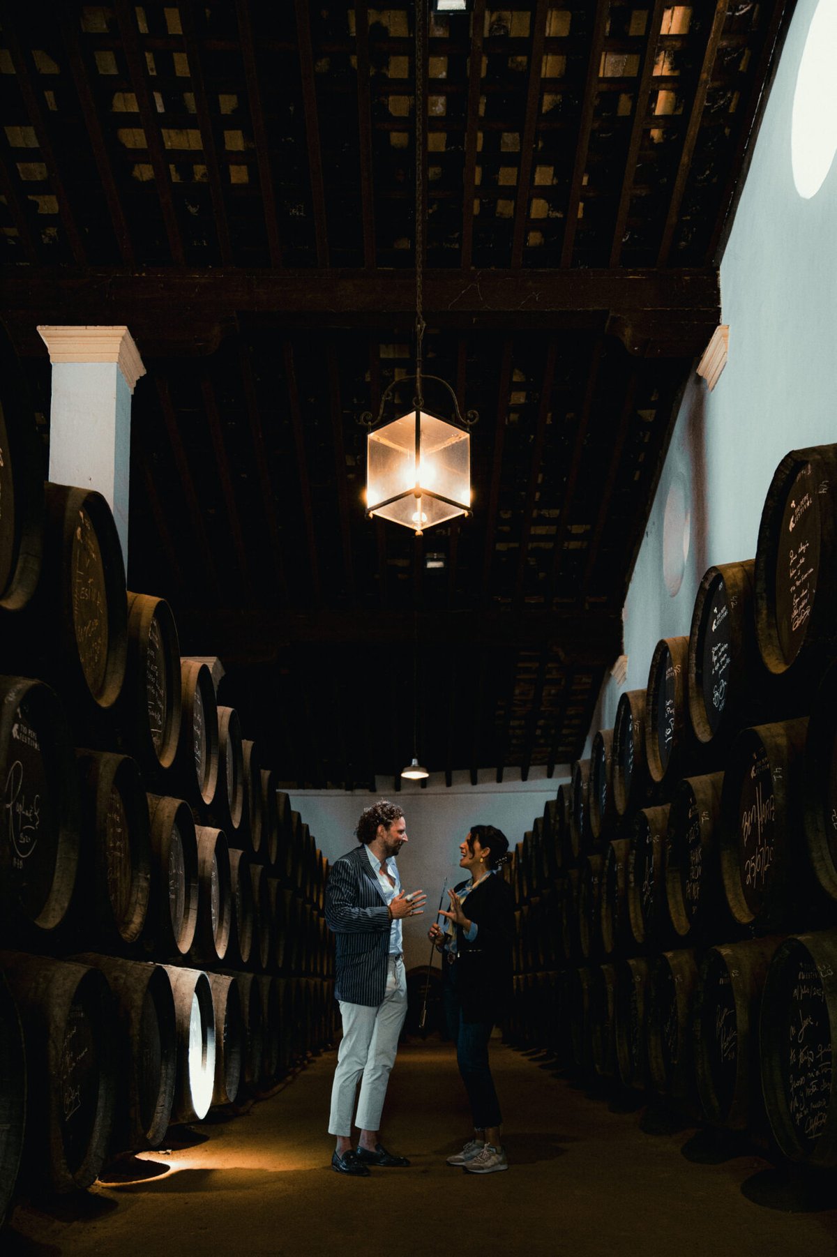 Twee mensen in gesprek in een sherrybodega tussen hoog opgestapelde rijen oude eikenhouten vaten