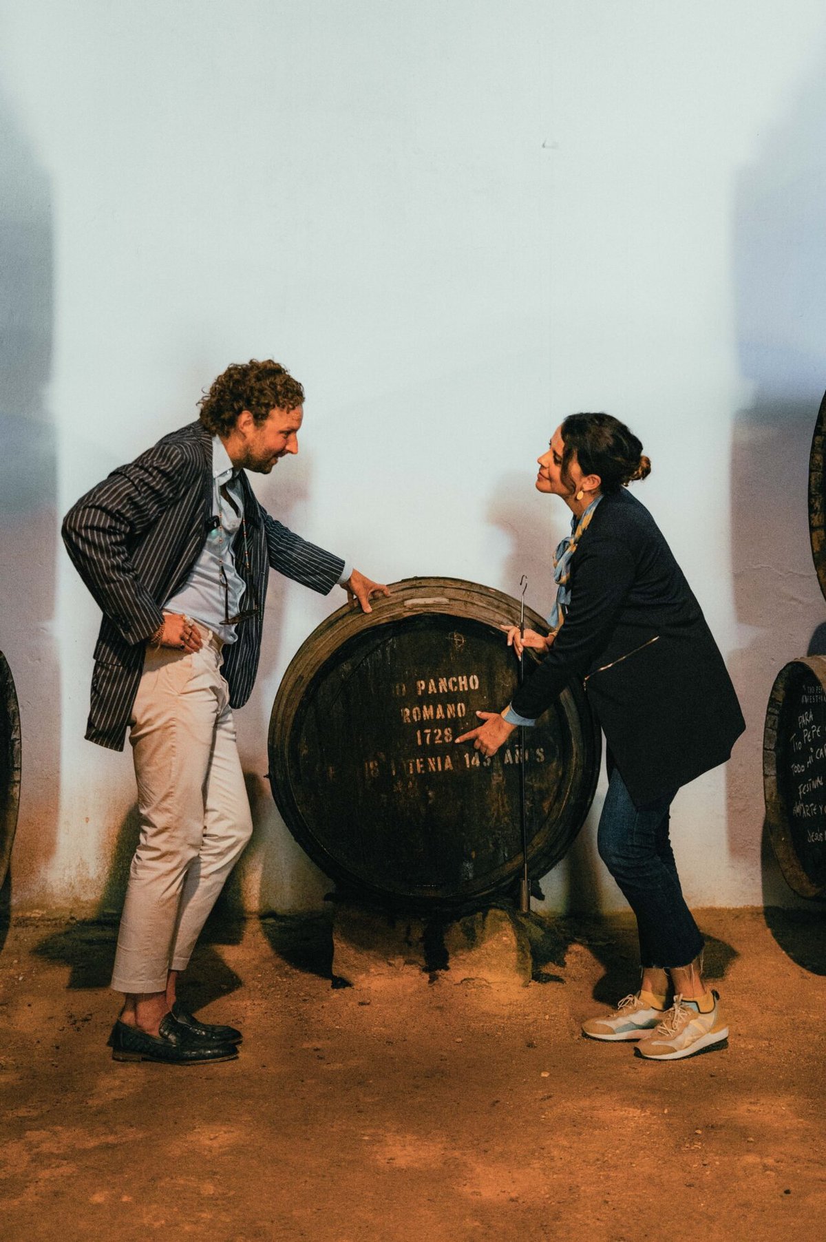 Een man en vrouw buigen zich naar een groot oud sherryvat met het opschrift Pancho Romano tegen een witte muur