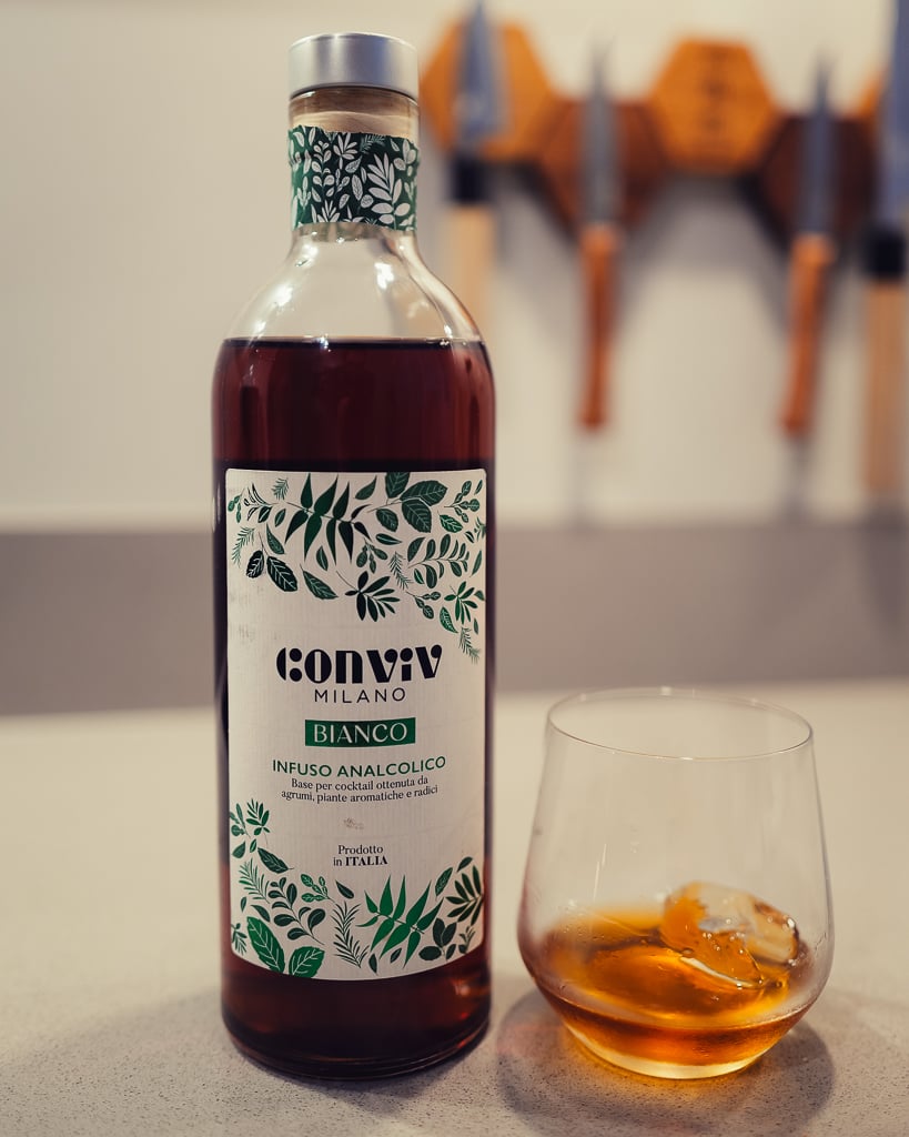 Fles Conviv Milano Bianco met zilveren dop naast een tumbler met amberkleurige drank en een ijsblokje