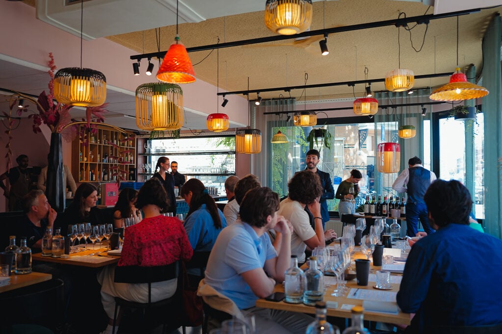 Druk restaurantinterieur met gasten aan tafels vol wijnglazen, kleurrijke hanglampen en een bar op de achtergrond