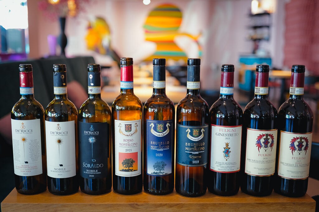 Rij van negen Toscaanse wijnflessen, waaronder De' Ricci en Fuligni, op een tafel in een kleurrijk restaurant