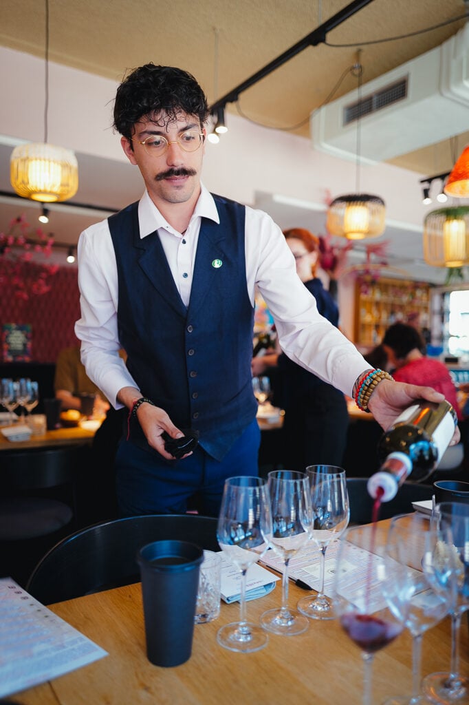 Een sommelier met snor en bril schenkt rode wijn in glazen aan een tafel in het restaurant