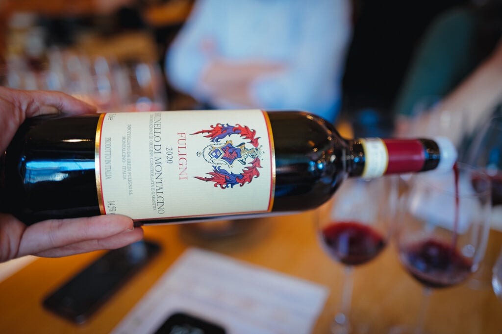 Een hand houdt een liggende fles Fuligni Brunello di Montalcino 2020 vast boven een tafel met wijnglazen