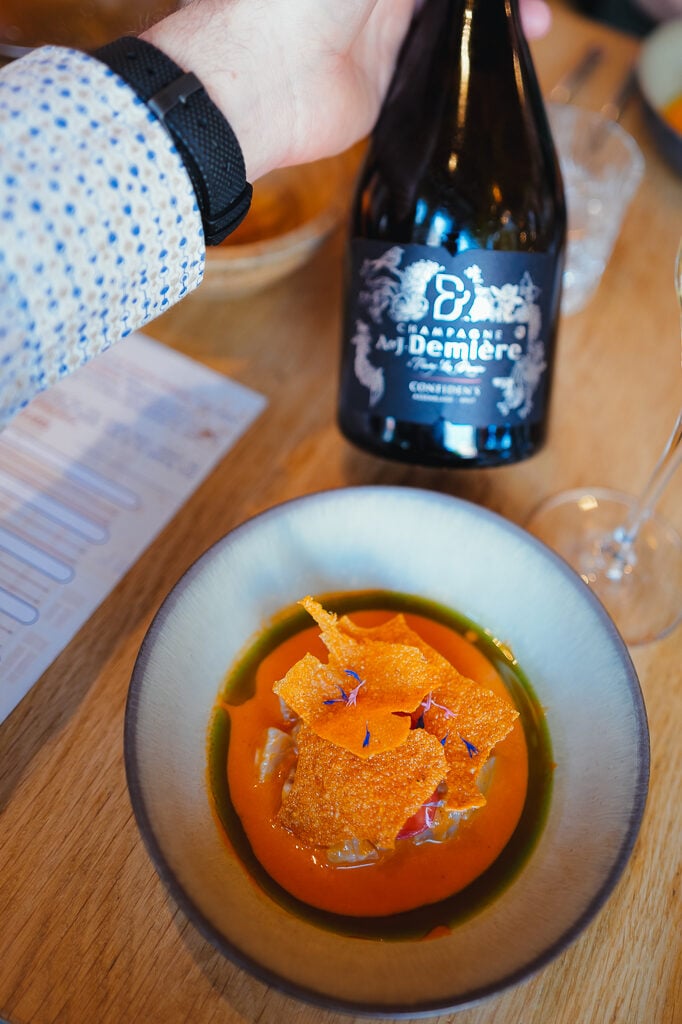 Een hand houdt een fles champagne bij een kom met romige oranje soep, krokante chips en eetbare bloemen