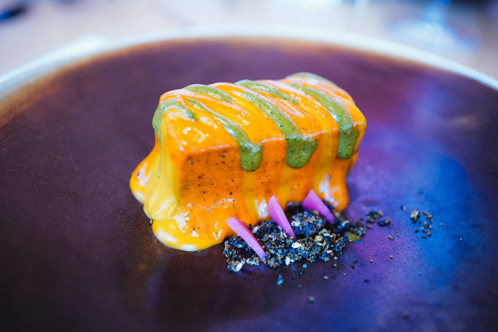 Verfijnd gerecht met oranje en groene saus en paarse uiringen op een donker bord