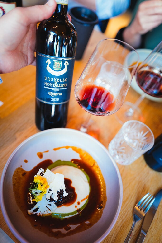 Een hand houdt een fles Brunello di Montalcino Riserva 2019 vast naast een glas rode wijn en een bord met saus