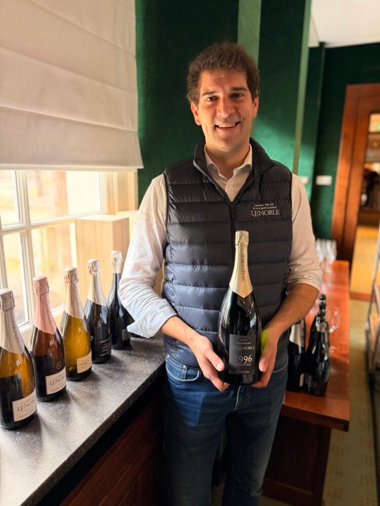 Glimlachende man in bodywarmer met AR Lenoble-logo houdt een fles champagne 1996 vast bij een rij flessen