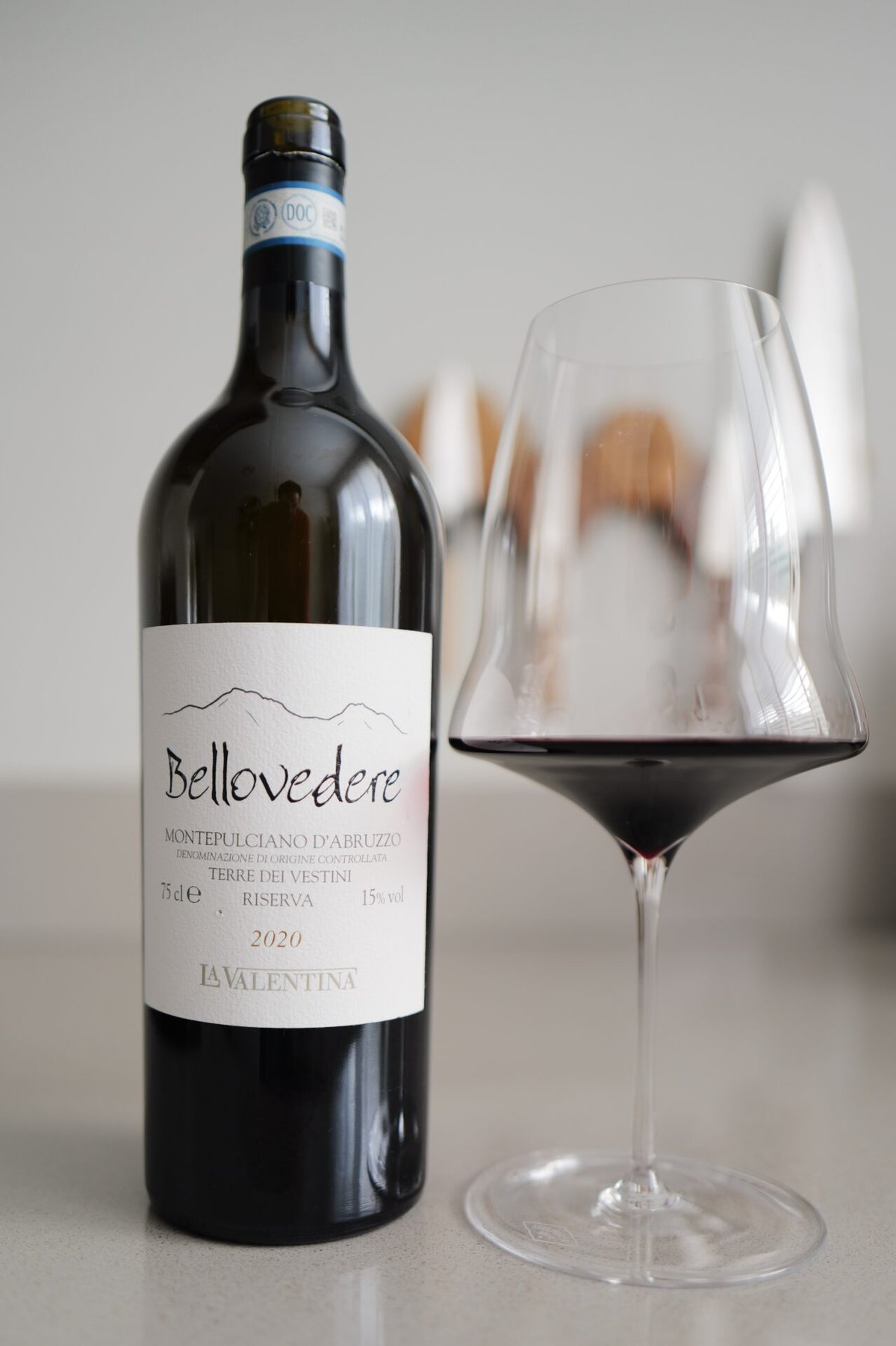 Fles Bellovedere 2020 Montepulciano d'Abruzzo van La Valentina naast een glas rode wijn op een lichte ondergrond