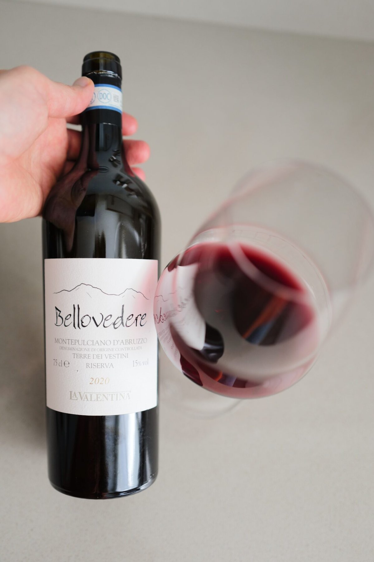 Een hand houdt een fles Bellovedere 2020 vast naast een gekanteld glas diepe rode wijn