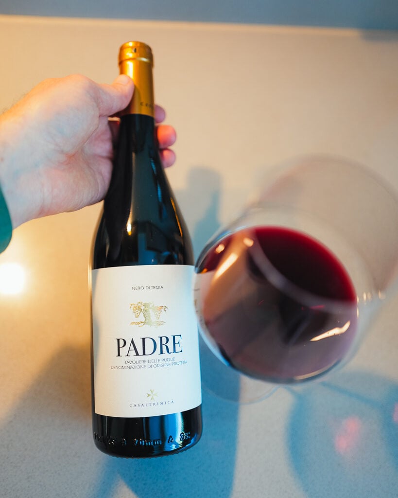 Een hand houdt een fles Padre Nero di Troia vast naast een gekanteld glas rode wijn