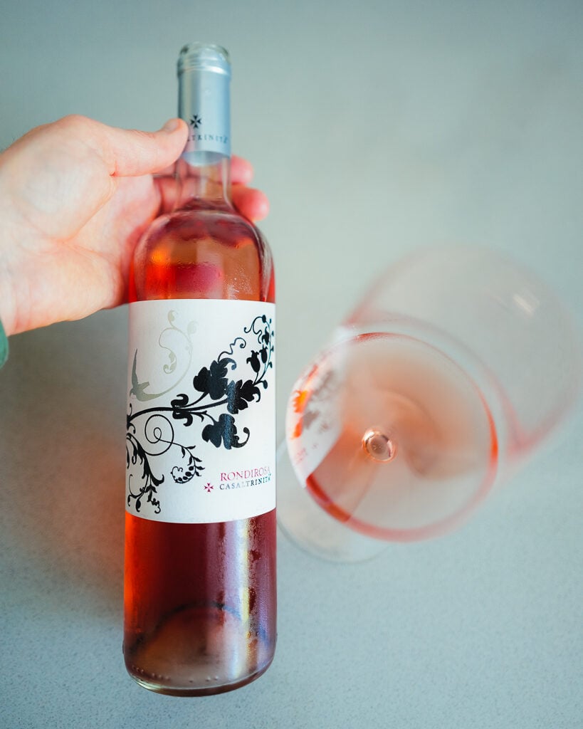 Een hand houdt een fles Rondirosa rosé vast naast een gekanteld glas zalmroze wijn