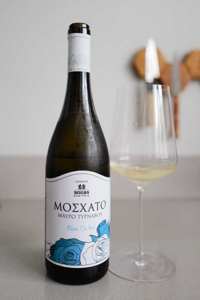 Fles Moschato Mavro Tirnavou Blanc de Noir met blauw rozenmotief-etiket naast een glas lichte witte wijn