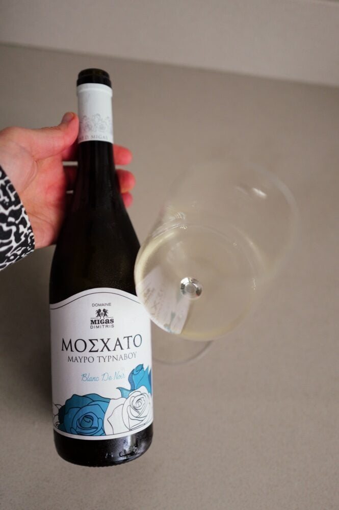 Een hand houdt een fles Moschato Mavro Tirnavou Blanc de Noir vast naast een gekanteld glas lichte witte wijn