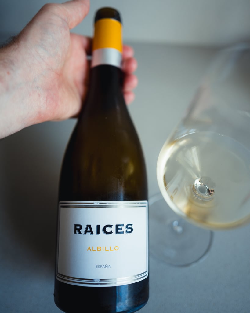 Hand houdt fles Raíces Albillo witte wijn vast bij een glas goudgele wijn