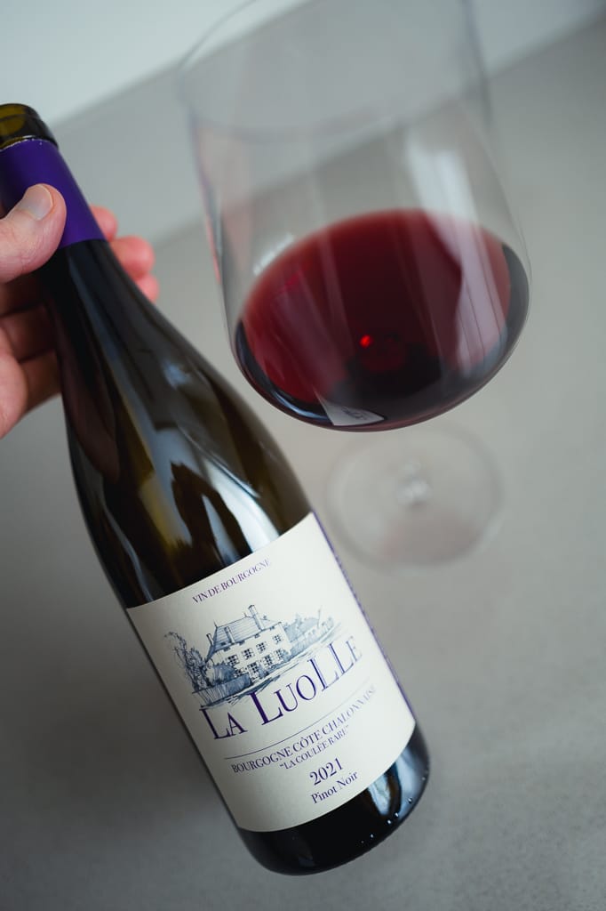 Hand houdt fles La Luolle Bourgogne Côte Chalonnaise Pinot Noir 2021 vast bij een glas rode wijn
