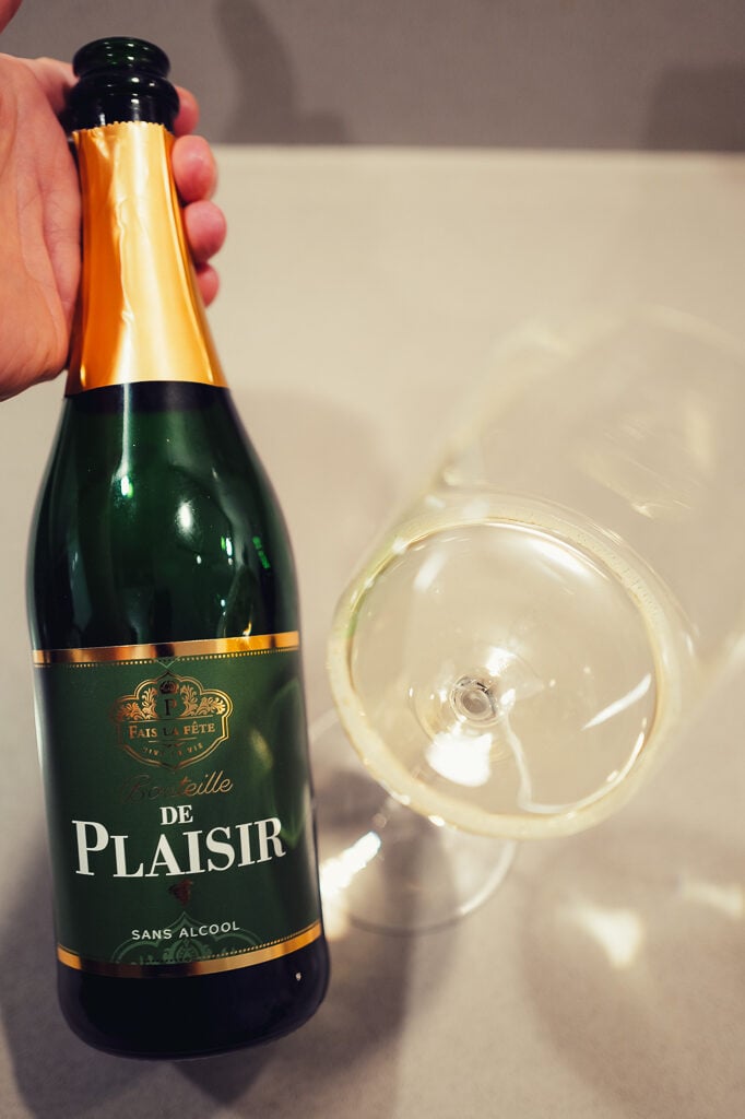 Hand houdt fles Bouteille de Plaisir sans alcool met groen-gouden etiket vast bij een glas