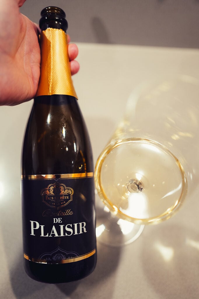 Hand houdt donkere fles Bouteille de Plaisir met goudfolie vast naast een glas mousserende wijn