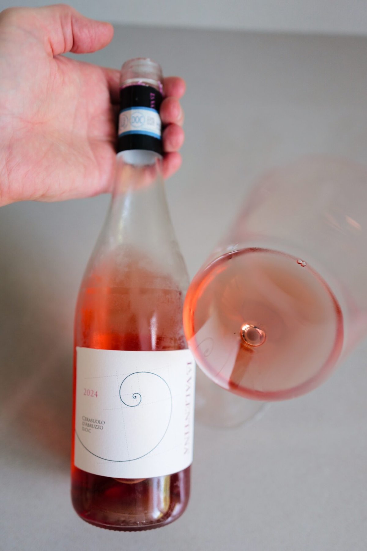 Hand houdt fles La Valentina Cerasuolo rosé vast naast een gekanteld glas met kersrode wijn