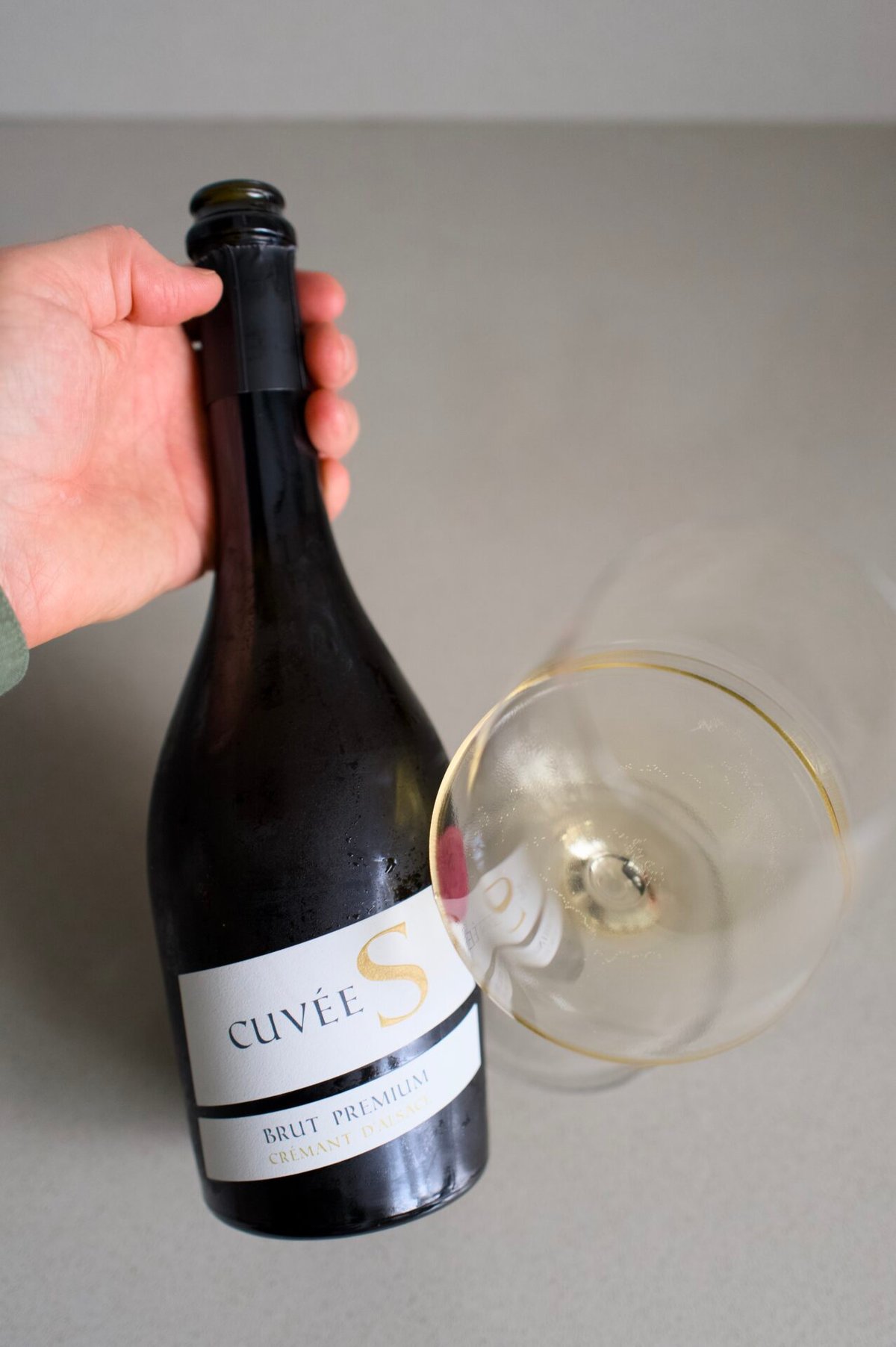 Hand houdt fles Cuvée S Crémant d'Alsace vast boven een glas mousserende wijn