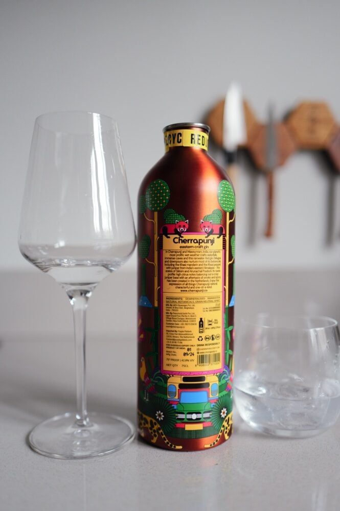 Achterkant van het geïllustreerde blik Cherrapunji gin met botanicals naast een wijnglas en tumbler