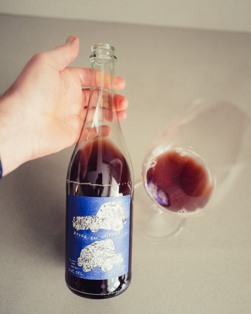 Hand holding the Cul Sec Rouge en Voiture bottle beside a glass of dark red drink