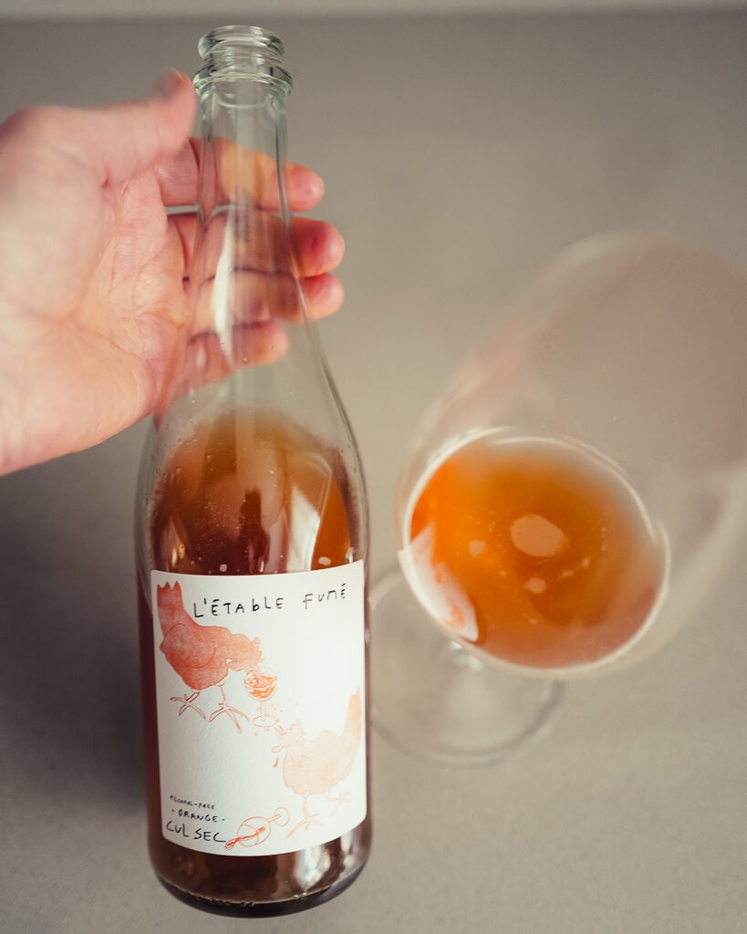 Hand holding the clear L'Étable Fumé bottle with hen illustration beside a glass of cloudy orange drink