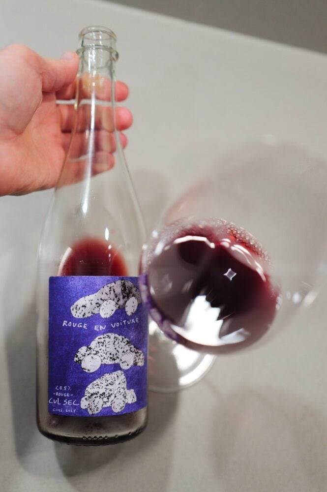 Hand houdt fles Cul Sec Rouge en Voiture vast naast een glas met donkerrode drank van bovenaf