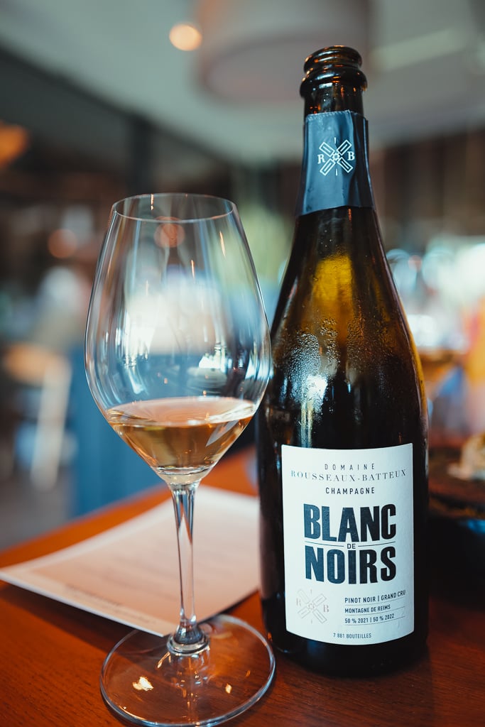 Fles Domaine Rousseaux-Batteux Blanc de Noirs champagne naast een glas op een houten tafel