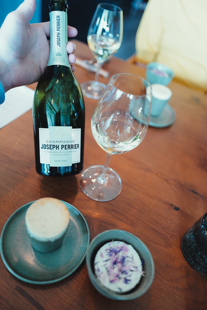 Hand houdt fles Champagne Joseph Perrier vast naast een glas en kleine amuses op een tafel