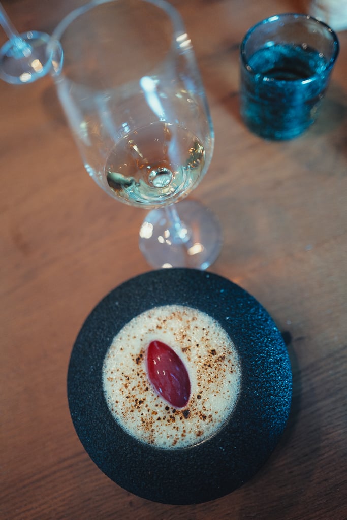 Dessertgerecht met schuim en een rode quenelle in een donkere kom naast een glas champagne