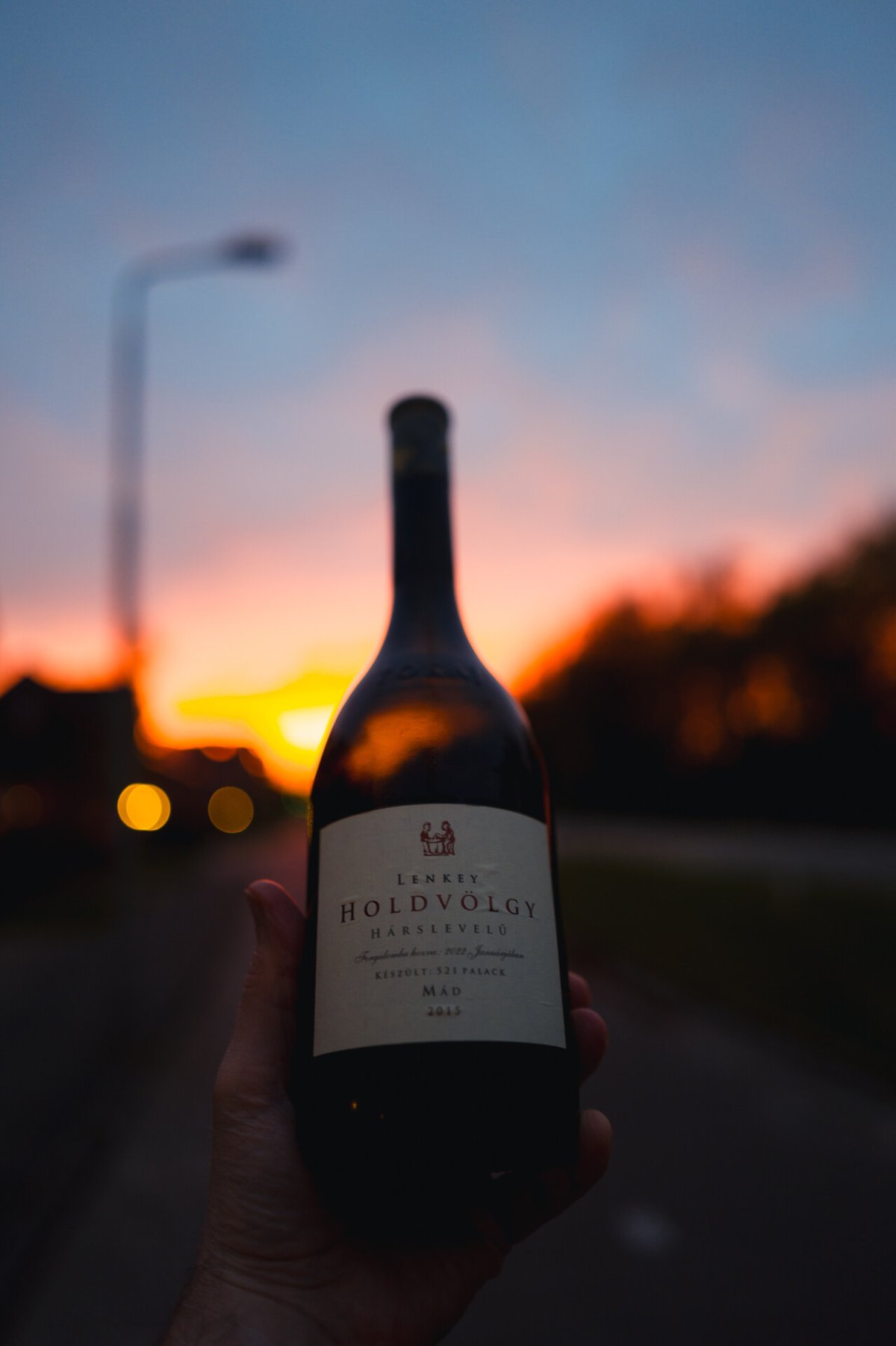 Hand houdt fles Lenkey Holdvölgy tegen een oranje zonsondergang boven een straat