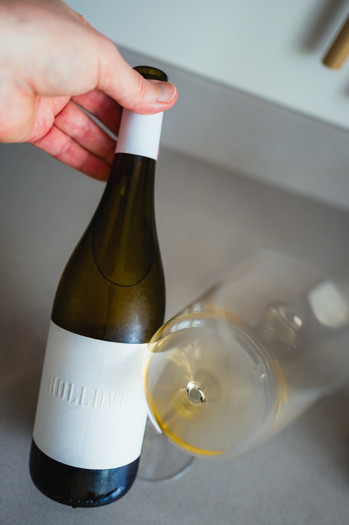 Hand houdt blanco Hollóvár-fles vast naast een liggend glas goudgele wijn