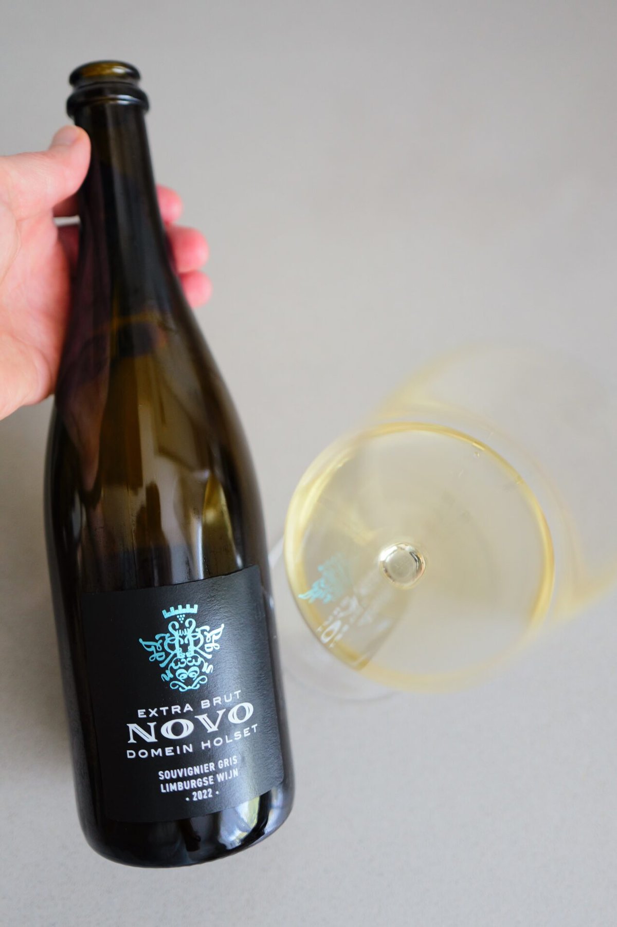Hand houdt fles Domein Holset Novo vast naast een liggend glas goudgele mousserende wijn