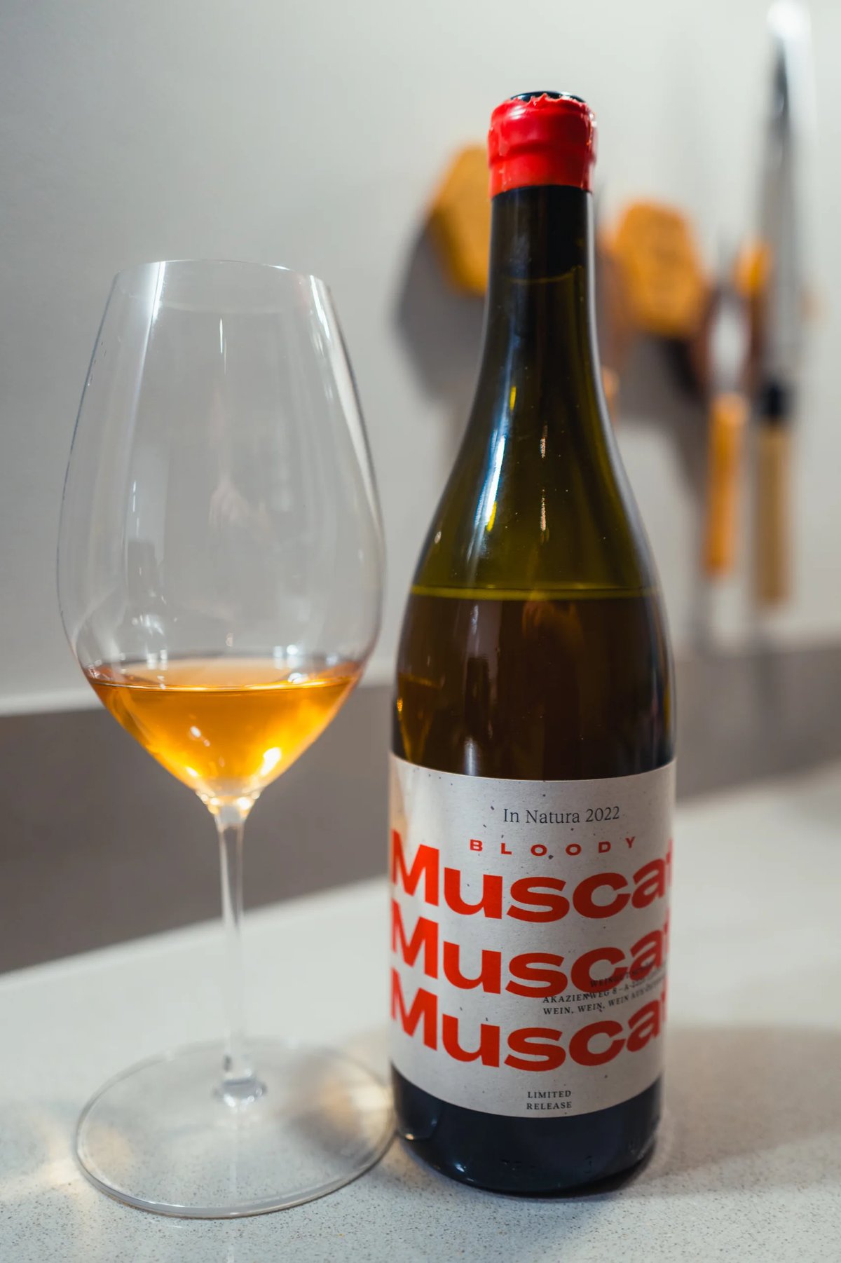 Fles Bloody Muscat In Natura 2022 met rood kapsel naast glas oranjewijn