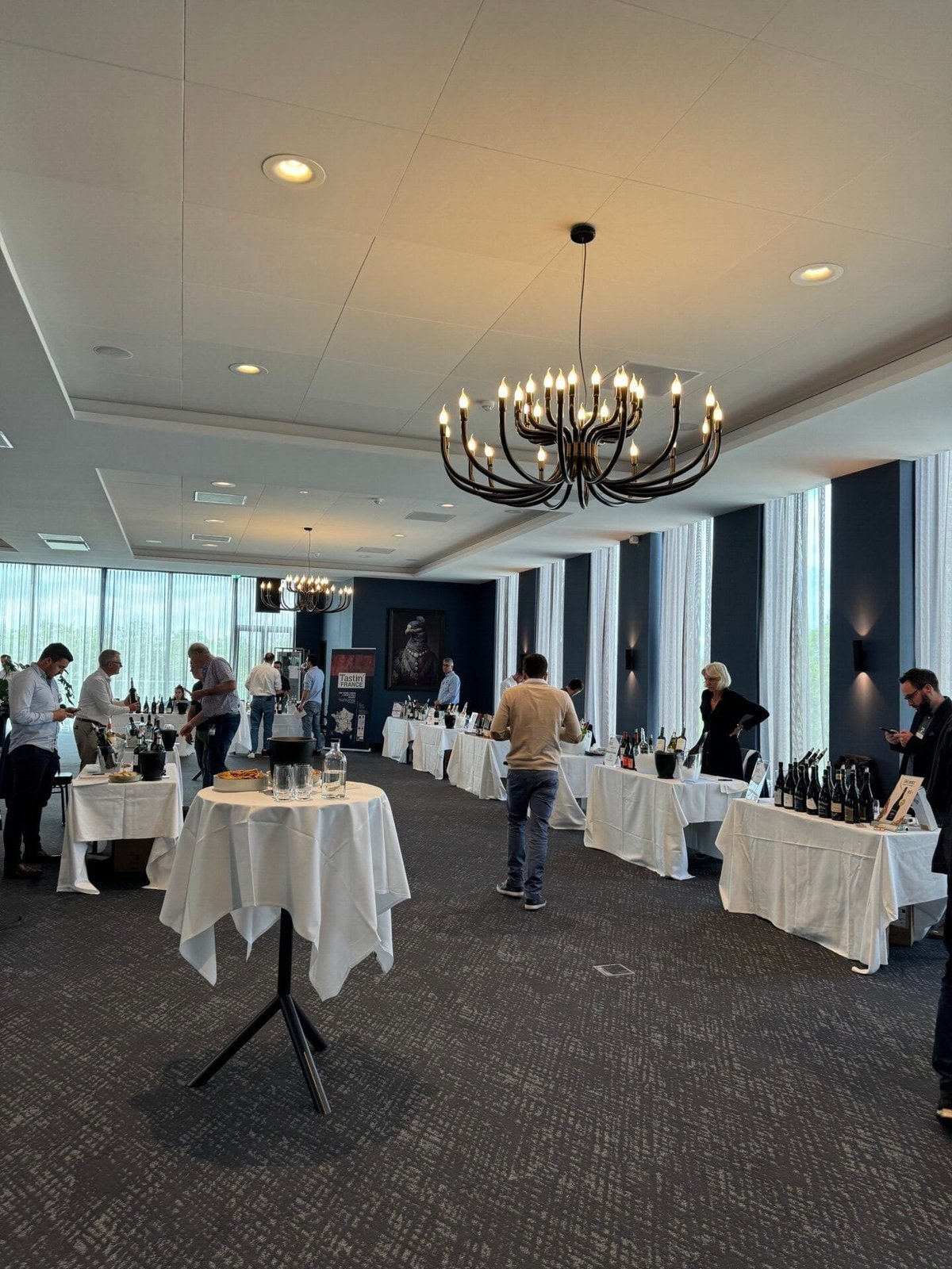 Proeverijzaal met statafels, witte tafelkleden, flessen en bezoekers onder kroonluchters