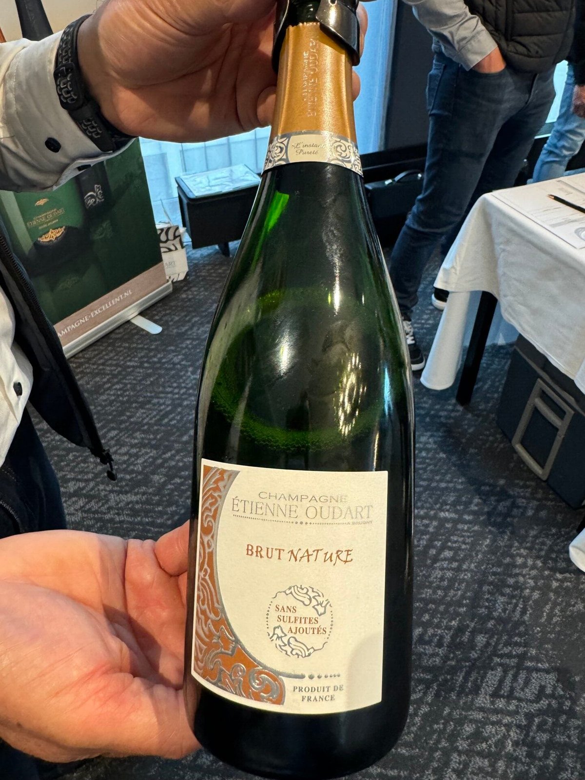 Hand houdt fles Champagne Étienne Oudart Brut Nature sans sulfites ajoutés vast op een beurs