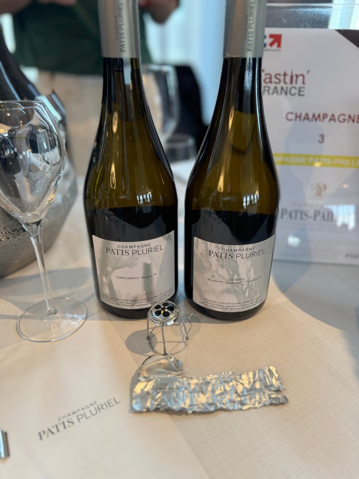 Twee flessen Champagne Patis Pluriel op een tafelkleed met champagnemuselet ervoor